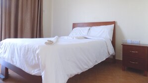 A neatly made bed with white linens, a wooden headboard, and a small wooden nightstand with drawers. - Plain Springs Hotel (Kumi)