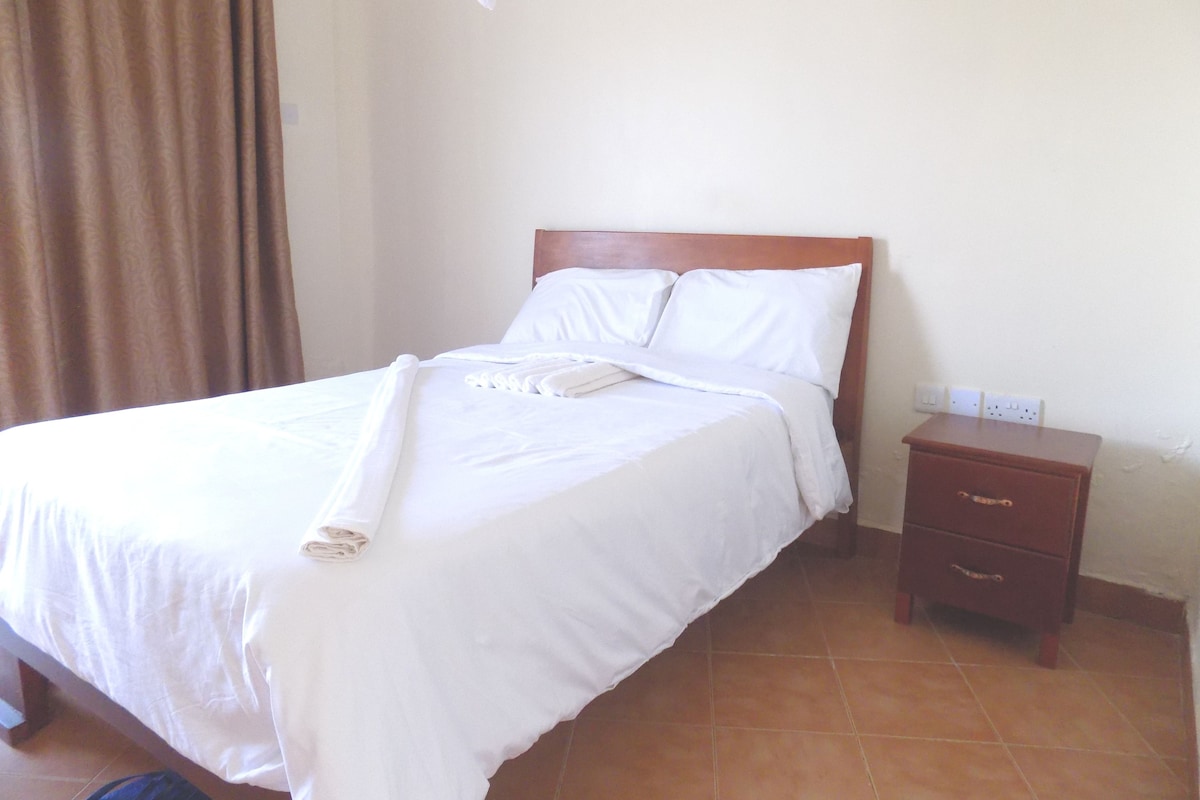 Una cama bien hecha con sábanas blancas y una toalla doblada, una mesita de noche de madera con un cajón, y una cortina marrón entreabierta.