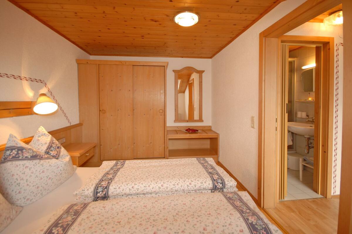A bedroom with a wooden ceiling, a bed with floral bedding, a bedside table, a mirror, and a wooden wardrobe.