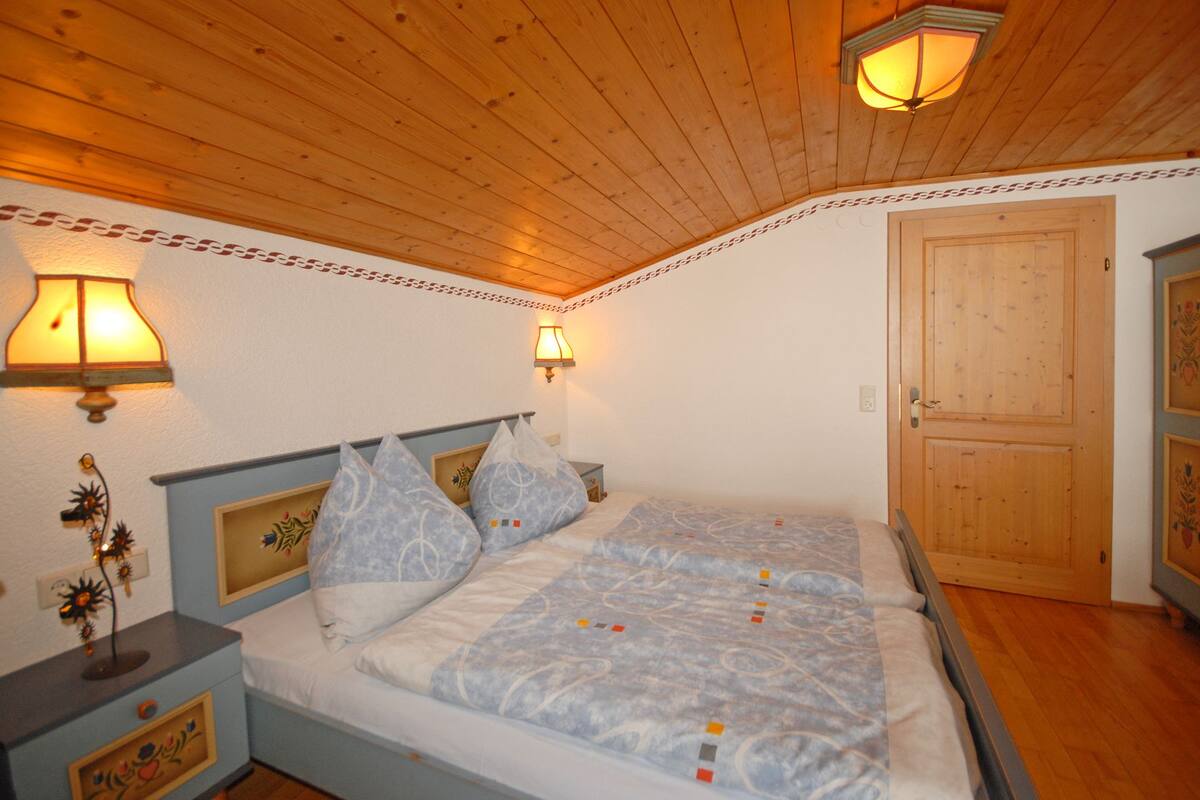 A bedroom with a wooden ceiling, two bedside lamps, a bed with patterned bedding, and a wooden door.