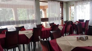 Una zona de menjador amb taules cobertes amb teles de taula vermelles i cadires, finestres grans amb cortines translúcides, i un sòl de rajola.