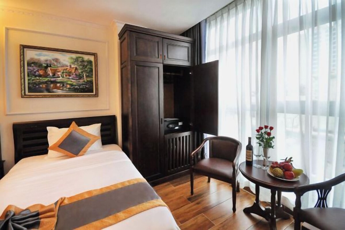 A hotel room with a bed, a dark wooden wardrobe, a desk with a chair, a small table with fruit and flowers, and a painting on the wall.