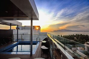 Un área de piscina en la azotea con barandilla de vidrio, zona de estar y vista al mar al atardecer.