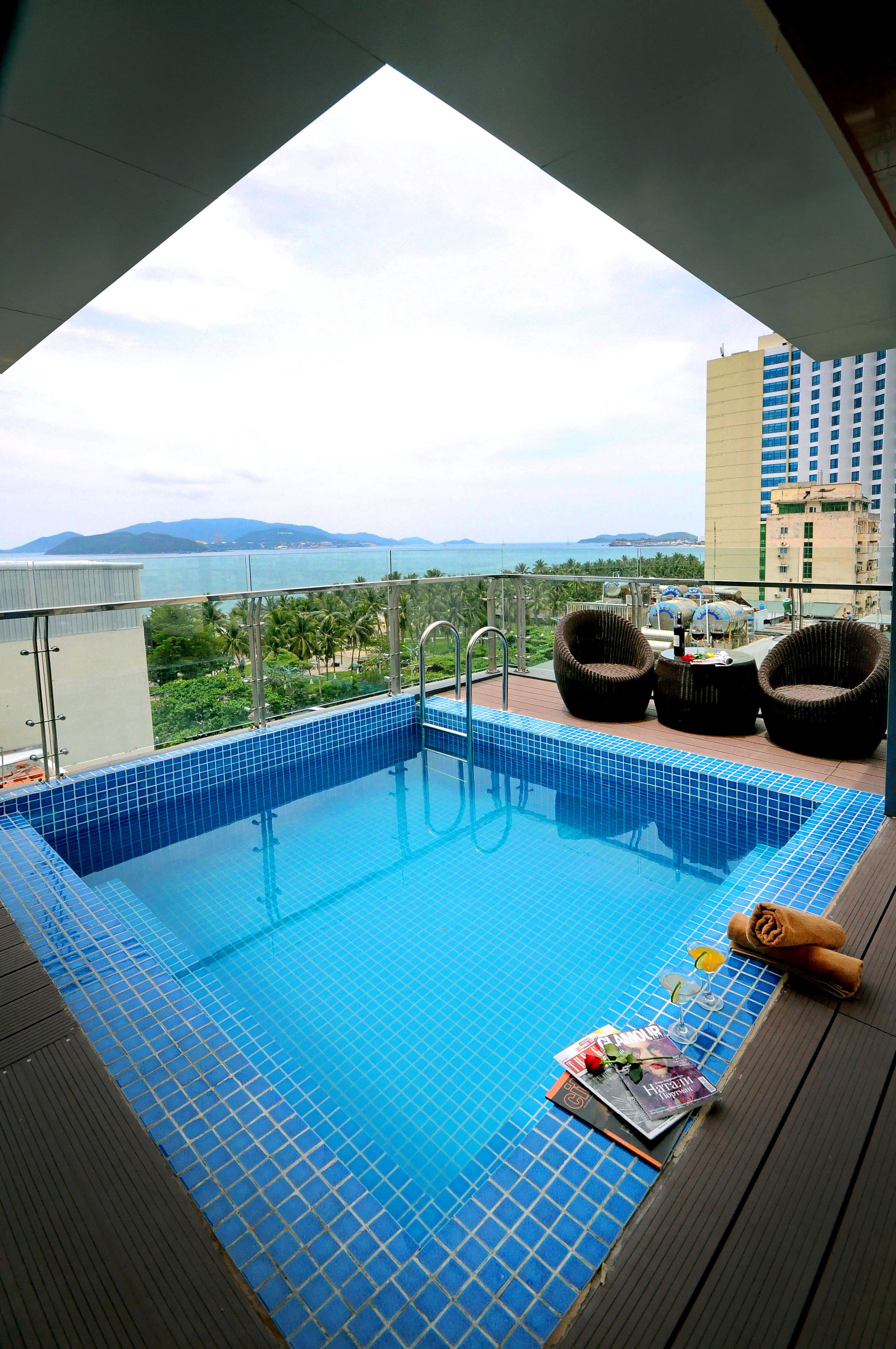 A rooftop swimming pool with a view of the sea and mountains, surrounded by wicker chairs and a glass railing.