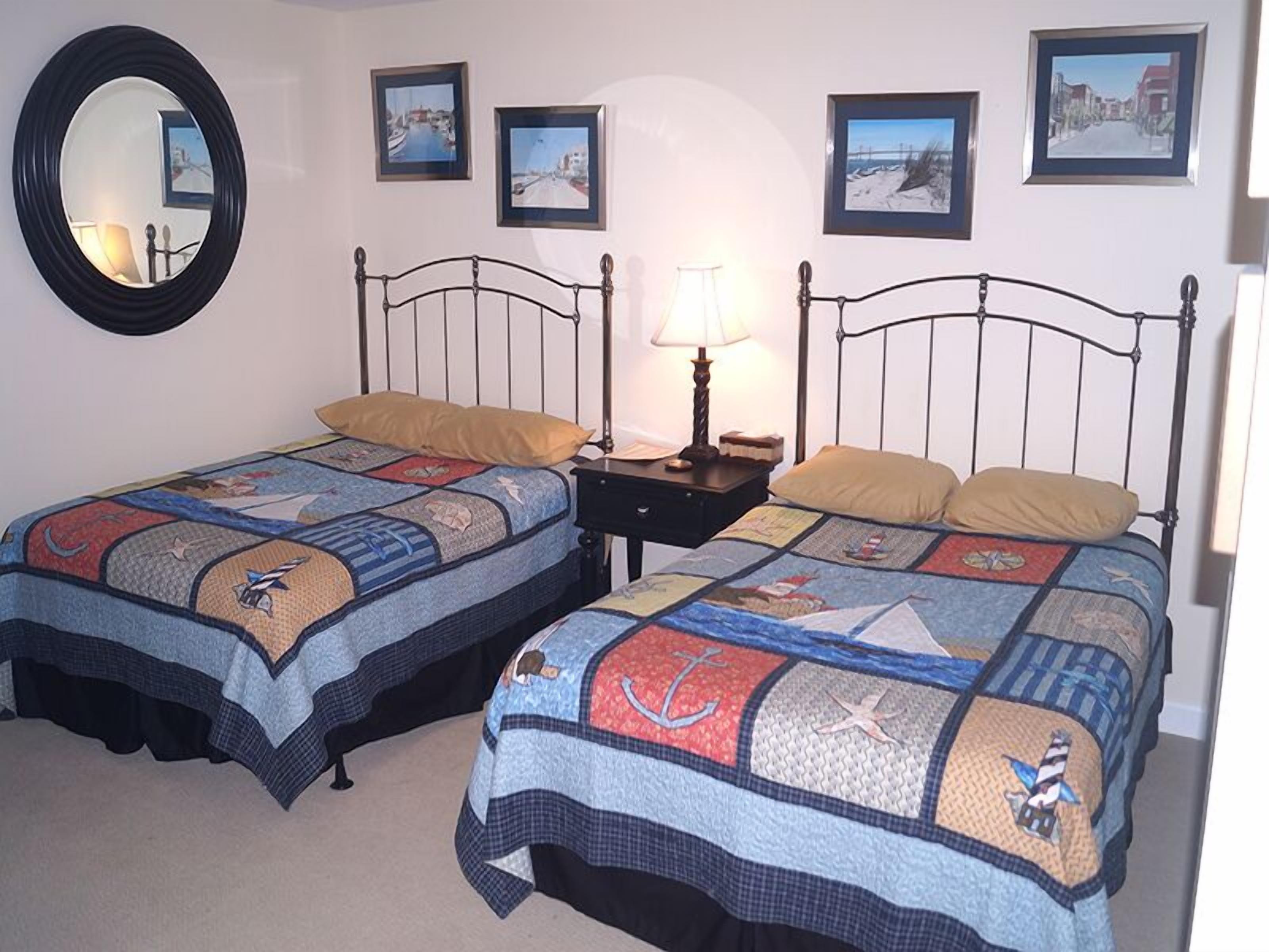 A room with two beds, each with a quilt featuring nautical themes. There are framed pictures on the wall, a round mirror, and a nightstand with a lamp.