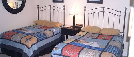 A room with two beds, each with a quilt featuring nautical themes. There are framed pictures on the wall, a round mirror, and a nightstand with a lamp.