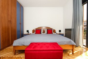 A bedroom with a wooden bed, red and grey bedding, a red bench, wooden wardrobe, and a view of the outside. - Apartment Tina with Hot Tub (Porec)