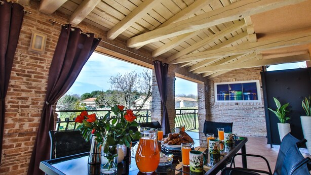 Une terrasse extérieure avec un mur en briques, des poutres en bois au plafond et une table dressée avec des aliments pour le petit-déjeuner.