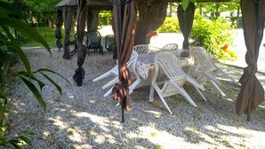 Uma área de estar coberta ao ar livre, com cadeiras de plástico branco e uma mesa, cercada por árvores e cascalho.