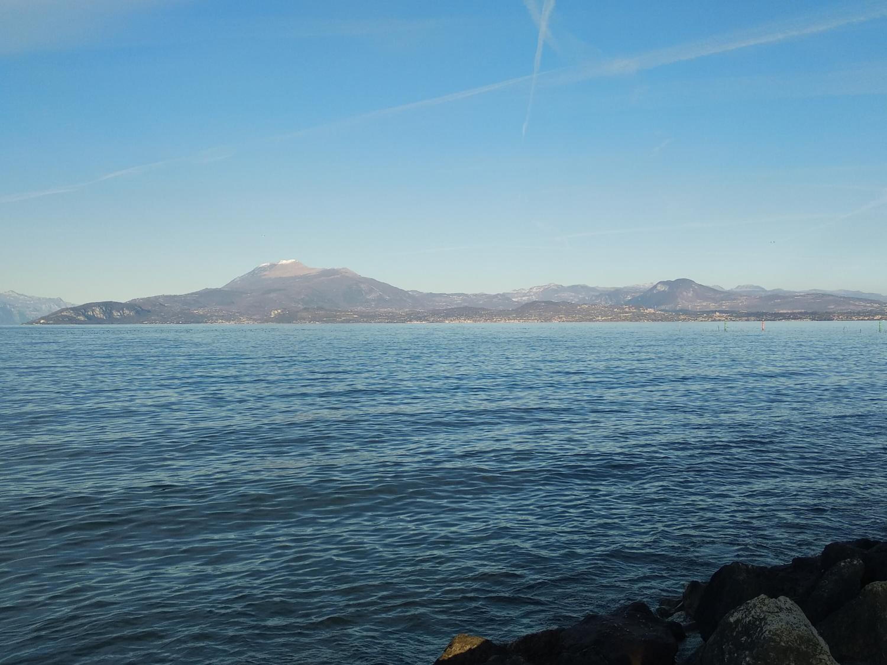 Paesaggio costiero con acque calme, costa rocciosa e montagne in lontananza.