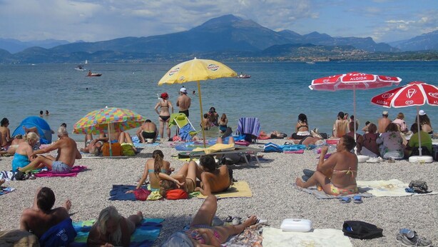 Praia cheia, com pessoas se banhando e tomando sol, guarda-sóis e barcos ao fundo.