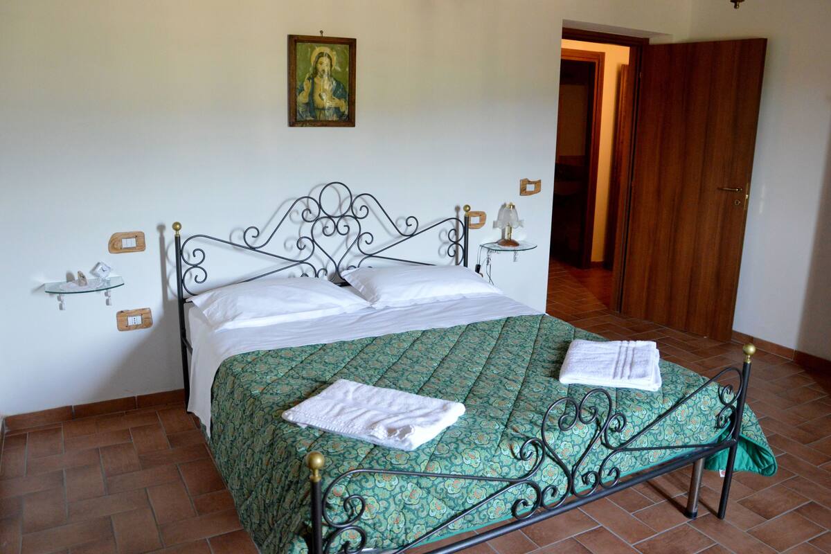 Un lit bien fait, recouvert d’une couvre-lit à motifs verts, avec deux serviettes blanches, une étagère fixée au mur sur laquelle repose un petit objet, et une porte donnant sur une autre pièce.