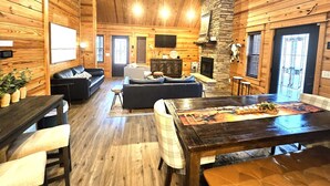 Ein gemütliches Blockhaus-Interieur mit Holzwänden, einem Kamin aus Stein, einer Essecke und einem Wohnzimmer mit Sofa und Sesseln.