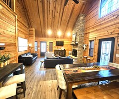 Interior acogedor de una cabaña con paredes de madera, una chimenea de piedra, un comedor y una sala de estar con sofá y sillones.