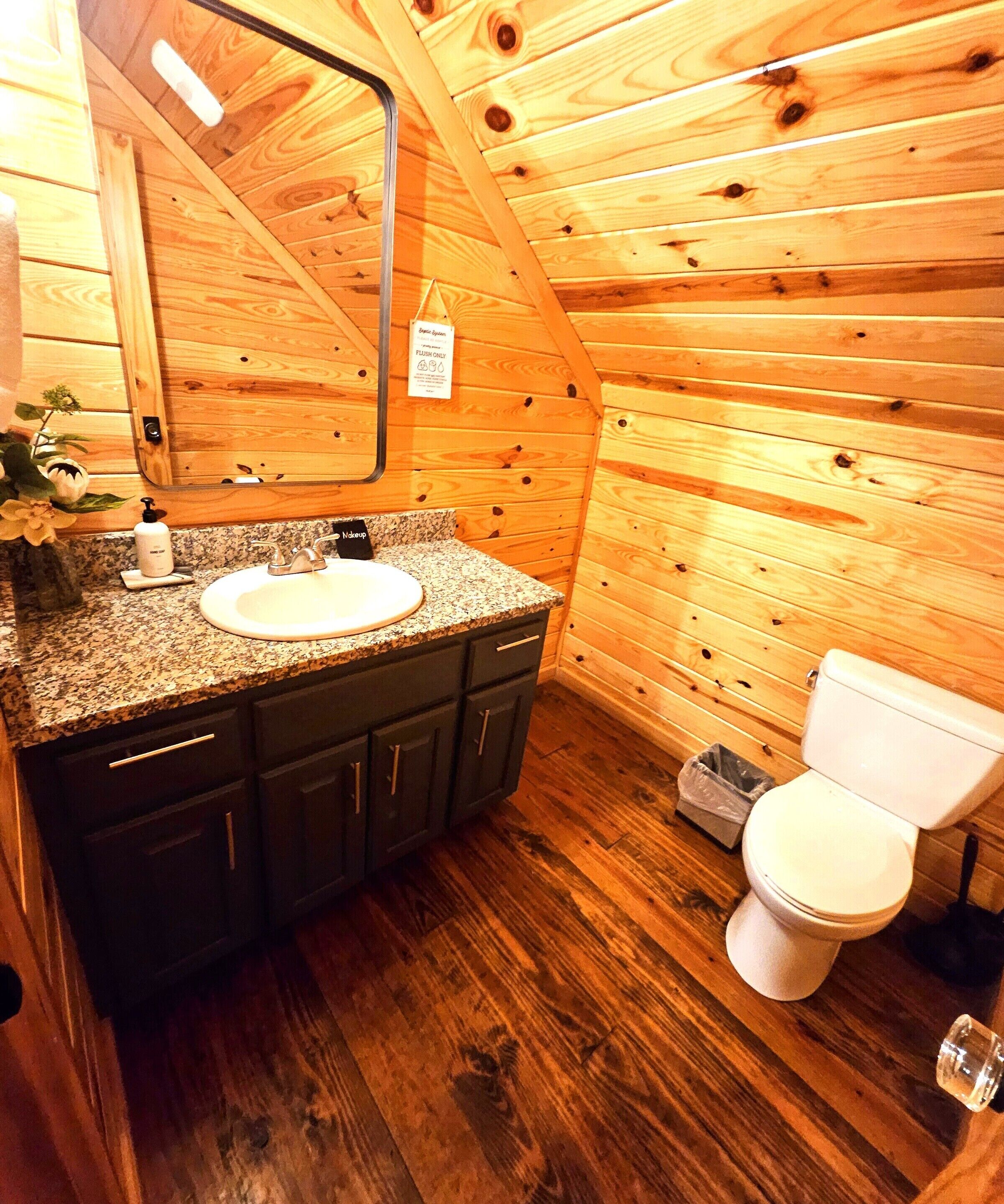 Un baño de cabaña de madera con lavabo de encimera de granito, armarios oscuros, inodoro blanco y espejo sobre el lavabo.