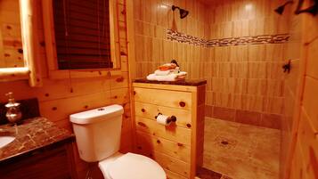 A wooden bathroom with a toilet, a wooden cabinet with a towel and a bottle, and a shower area with a tiled wall.