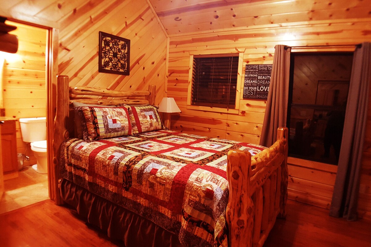 Ein gemütliches Schlafzimmer in einer Blockhütte mit einem hölzernen Bett, einem Nachttisch, einem Fenster mit Jalousien und einem an der Wand hängenden Bild.