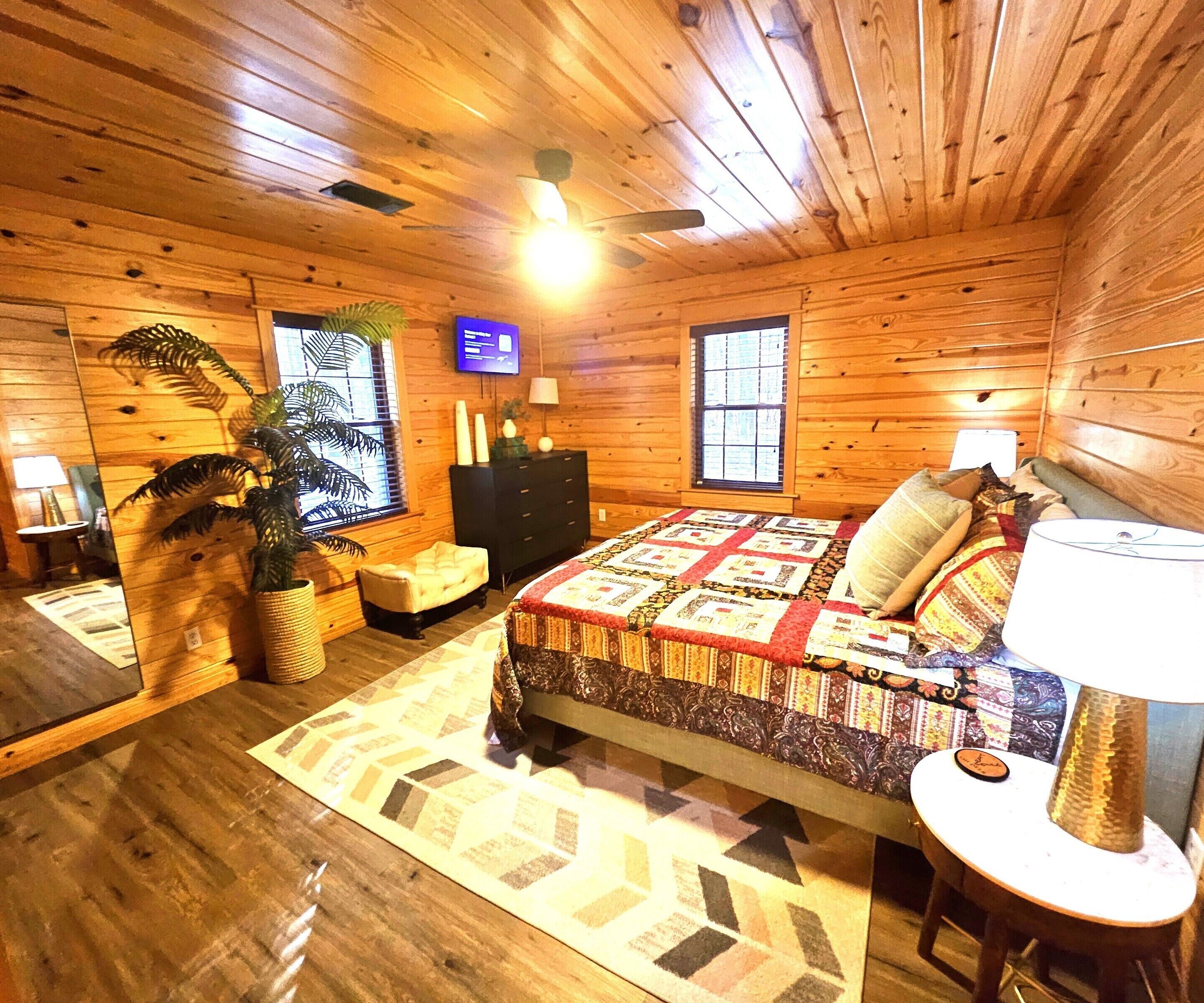 A cozy cabin-style room with a wooden interior, a bed with a patterned quilt, a television, and a window with blinds.