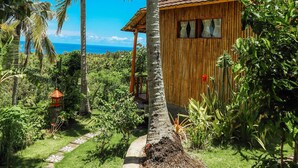 Un edificio de madera con techo de tejas, rodeado de exuberante vegetación y palmeras, con una vista despejada del océano al fondo.