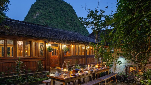 A wooden building with a tiled roof, a dining area with a long table and benches, and a view of a mountain in the background. - Yangshuo Secret Garden (Guilin)