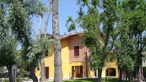 Un bâtiment jaune avec un toit en tuiles, entouré d’arbres et bordé d’une allée gravillonnée.
