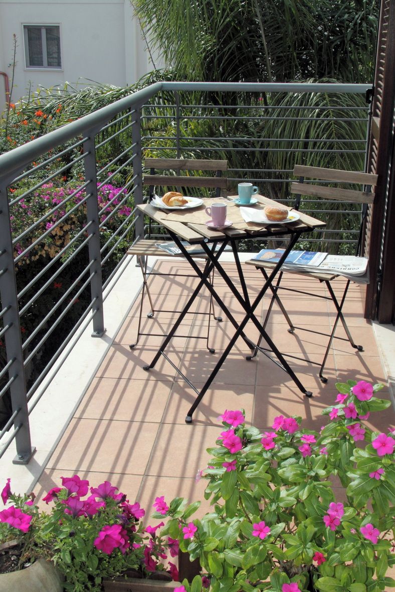 Un balcone con un tavolo apparecchiato per due persone, circondato da piante e fiori in vaso.