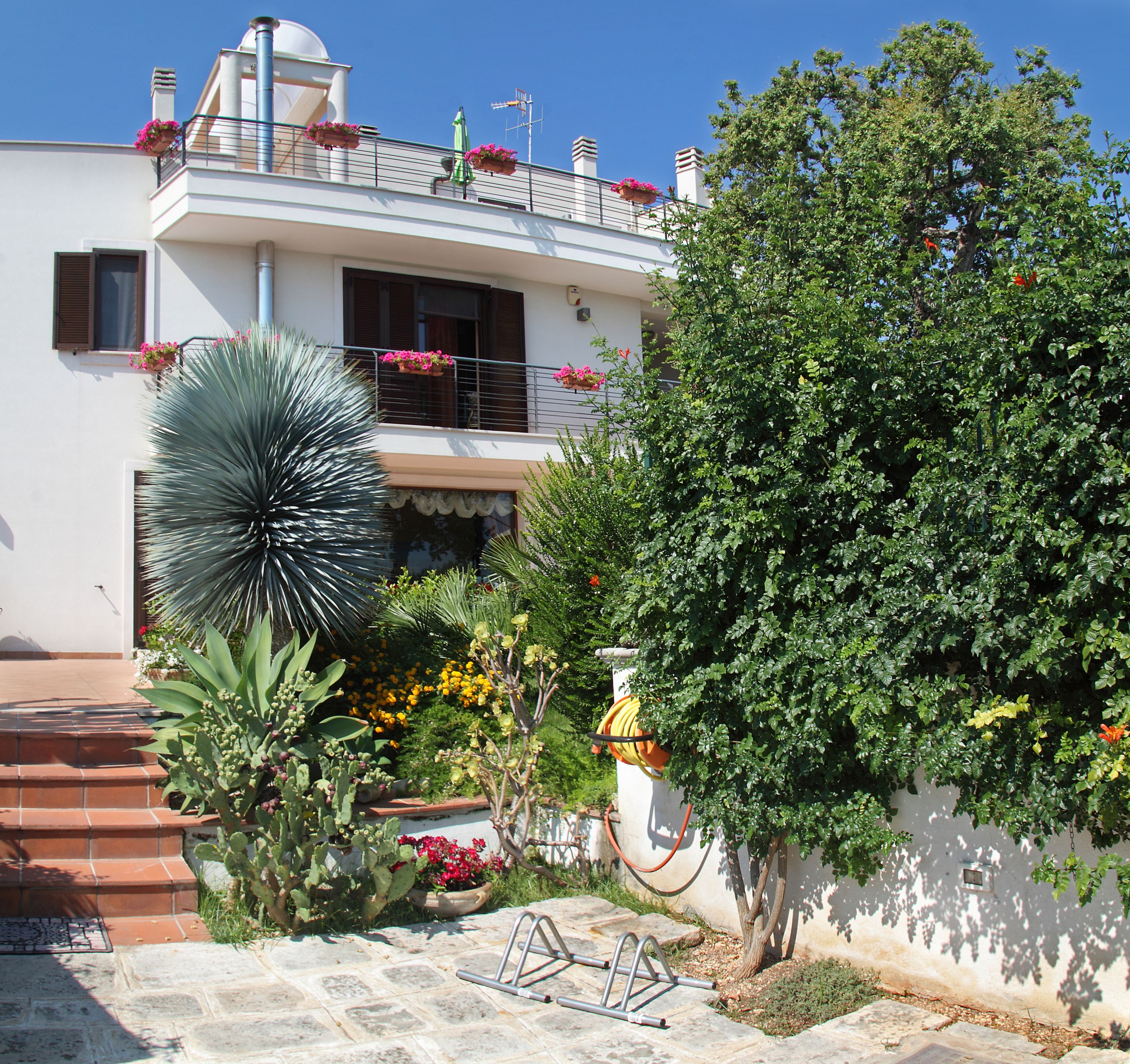 Un edificio a due piani con balconi, un giardino ricco di piante e fiori, e un cielo azzurro limpido.