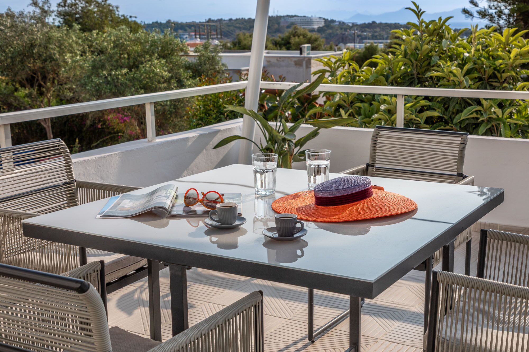 Un tavolo con un giornale, occhiali, un cappello e bicchieri d'acqua su un balcone con sedie e vista su spazi verdi ed edifici.