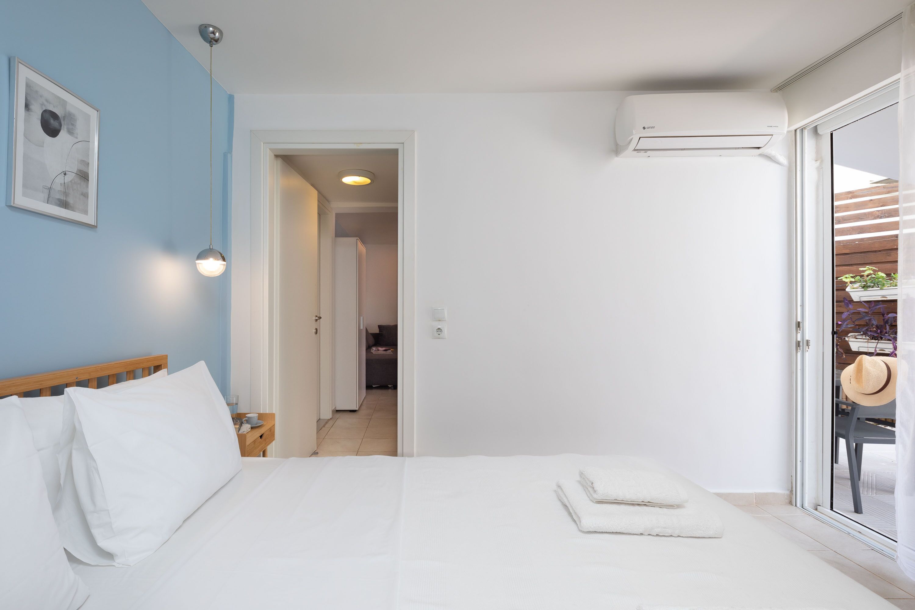 A bedroom with a bed, white bedding, a nightstand, a wall-mounted air conditioner, and a door leading to a balcony with a hat on it.