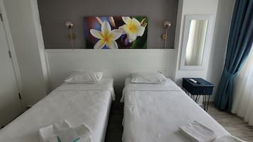A hotel room with two single beds, a bedside table, a nightstand, a painting of flowers, a mirror, and a window with curtains.