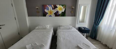 A hotel room with two single beds, a bedside table, a nightstand, a painting of flowers, a mirror, and a window with curtains.