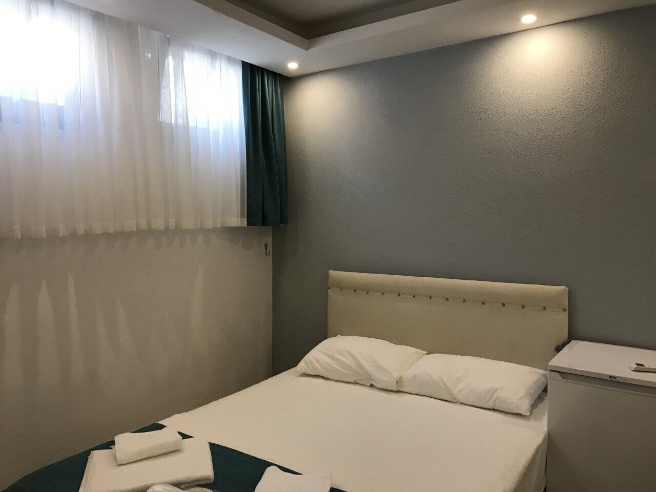 A clean, modern hotel room with a single bed, a bedside table, and a window with sheer curtains.