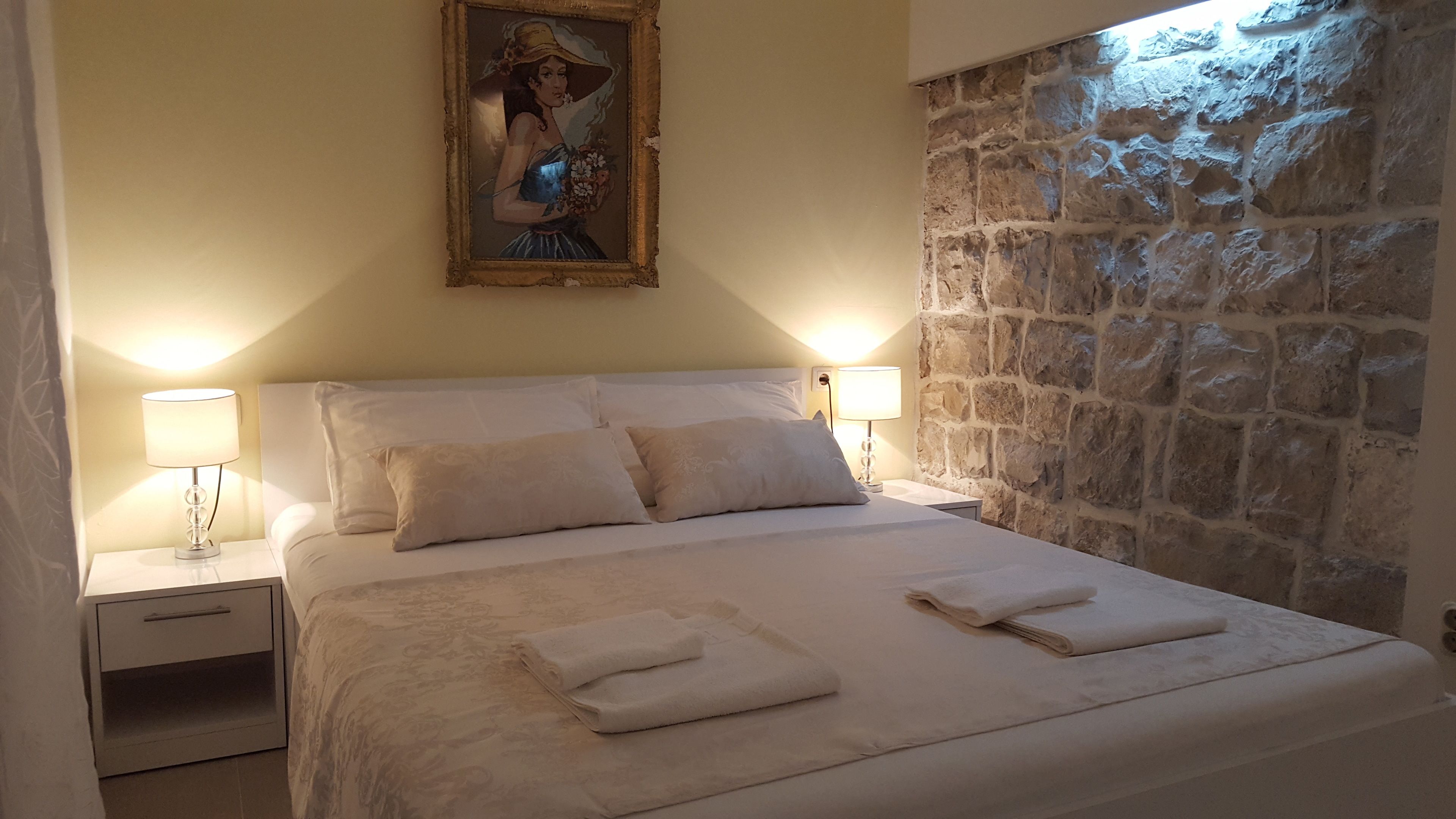 Een net opgemaakt bed met witte lakens en kussens, twee bedlampen, een schilderij boven het bed, en een stenen muur aan de rechterkant.