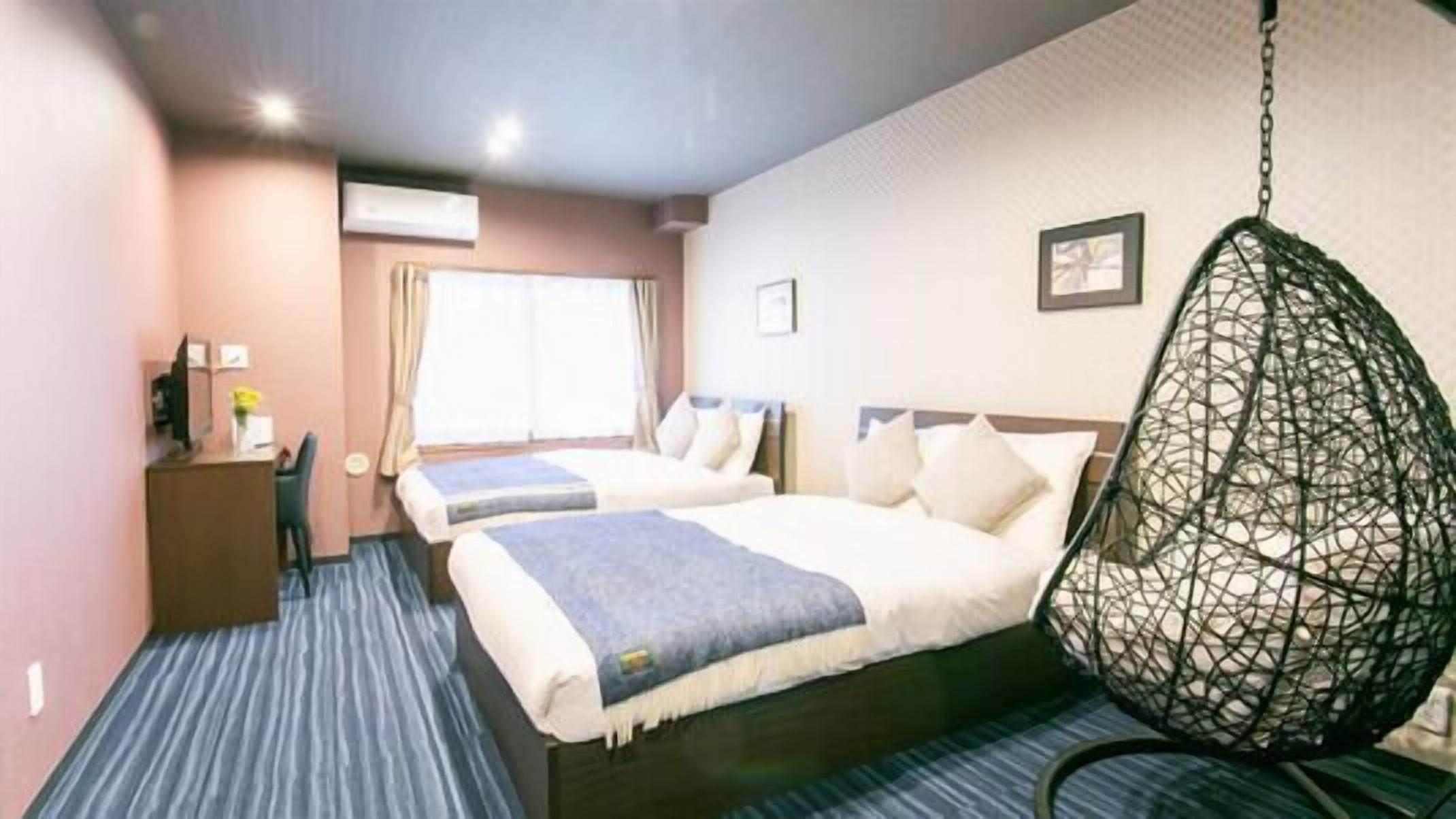 A hotel room with two beds, a desk, a chair, a hanging chair, and a window with curtains.