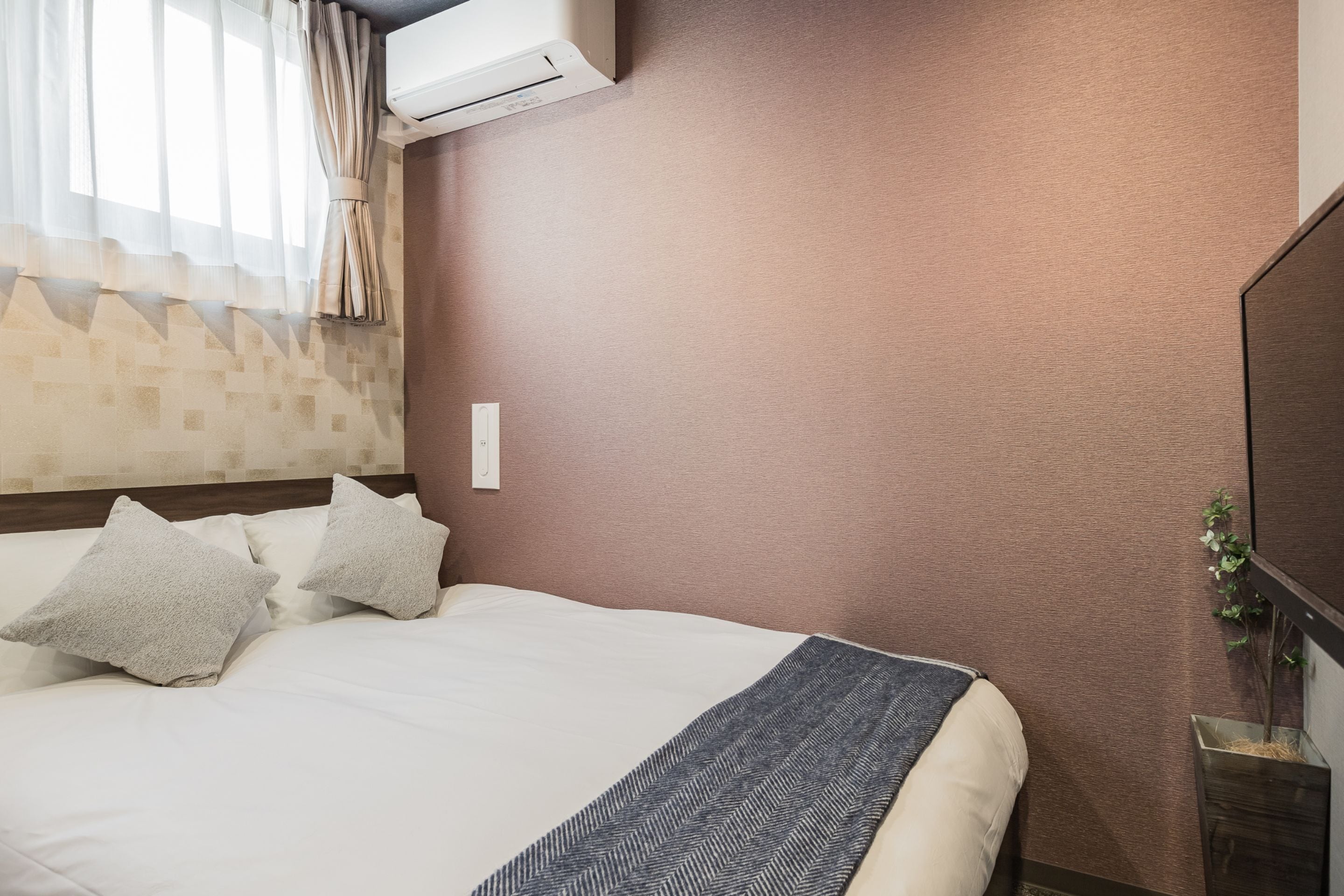 A hotel room with a bed, a wall-mounted air conditioner, a bedside table with a plant, and a window with sheer curtains.