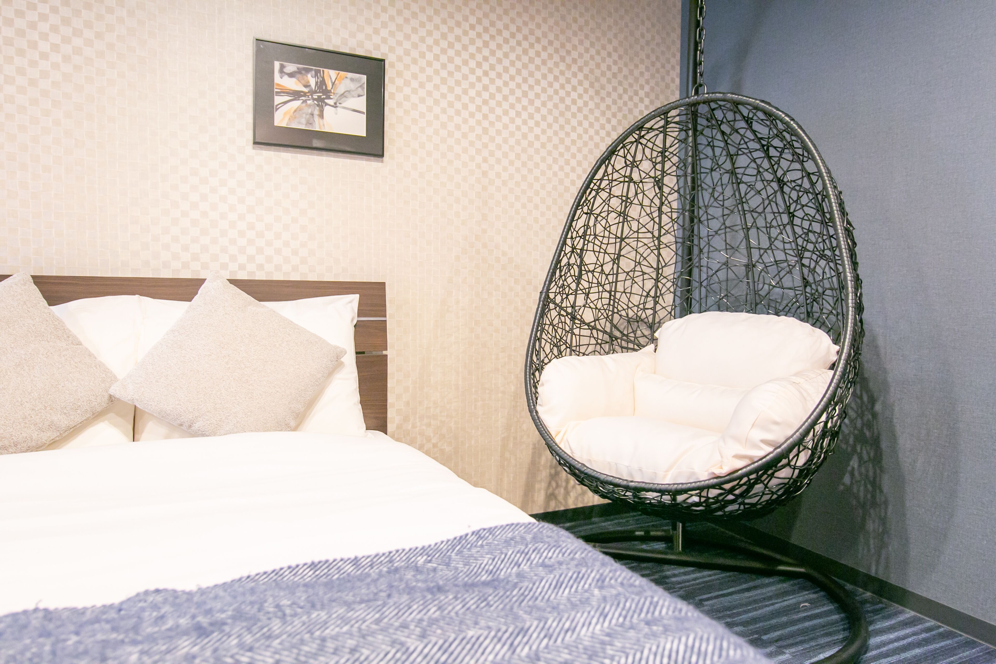A neatly made bed with white linens and two pillows, a hanging chair with a white cushion, and a framed picture on the wall.