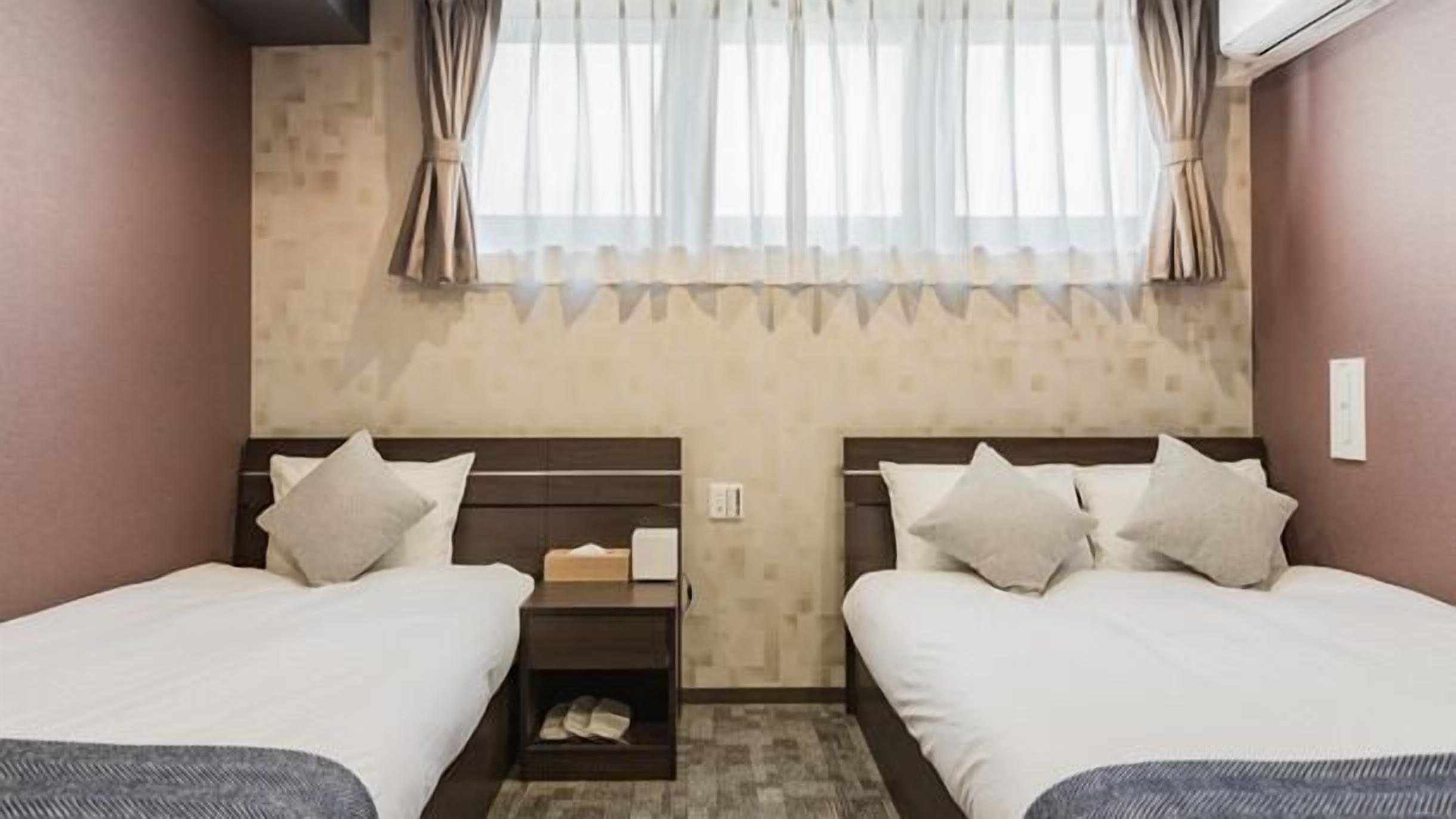 A hotel room with two beds, a nightstand with a lamp, a window with curtains, and a wall-mounted air conditioner.