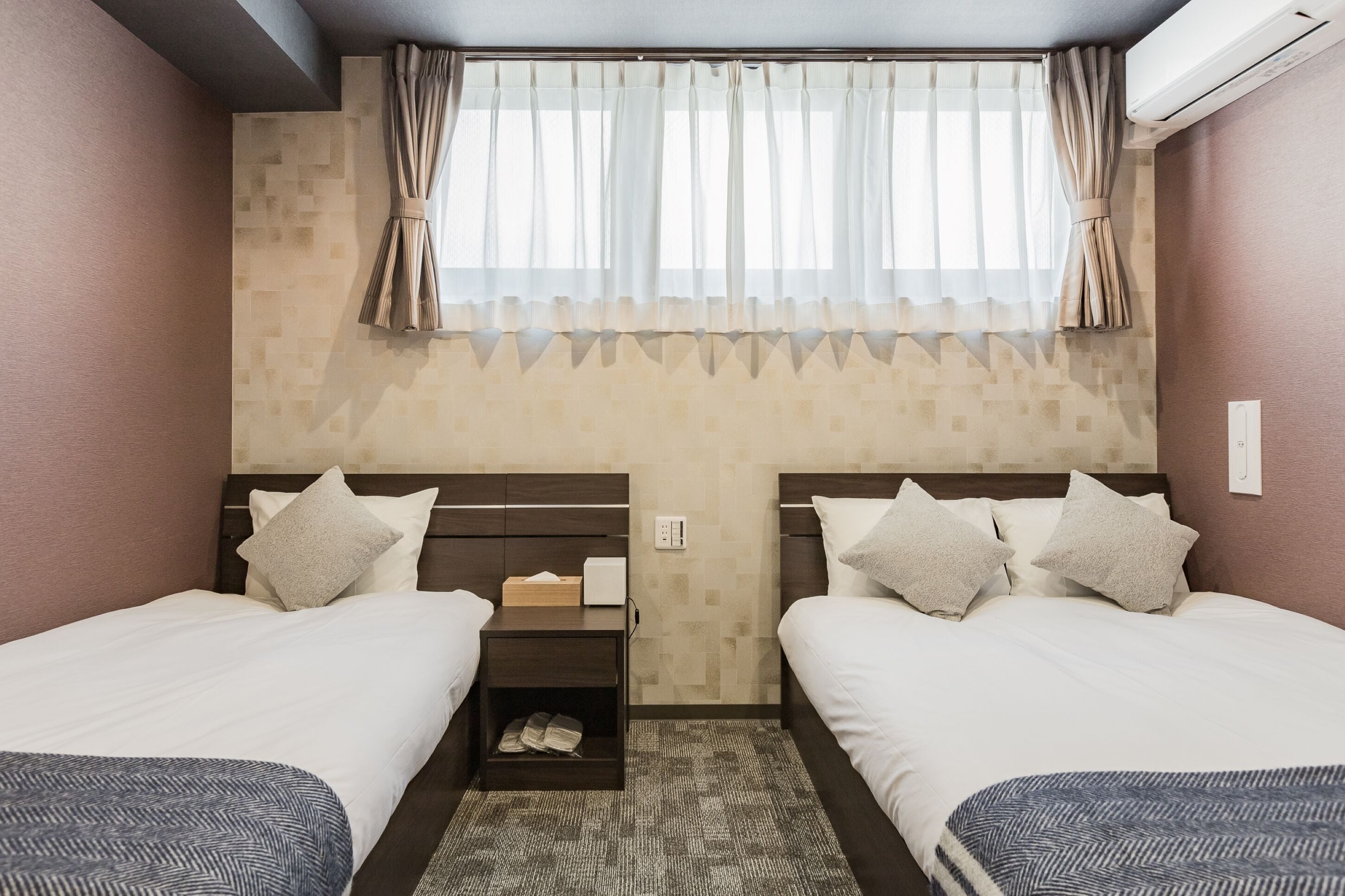 A hotel room with two beds, a nightstand, a small shelf, and a window with curtains.