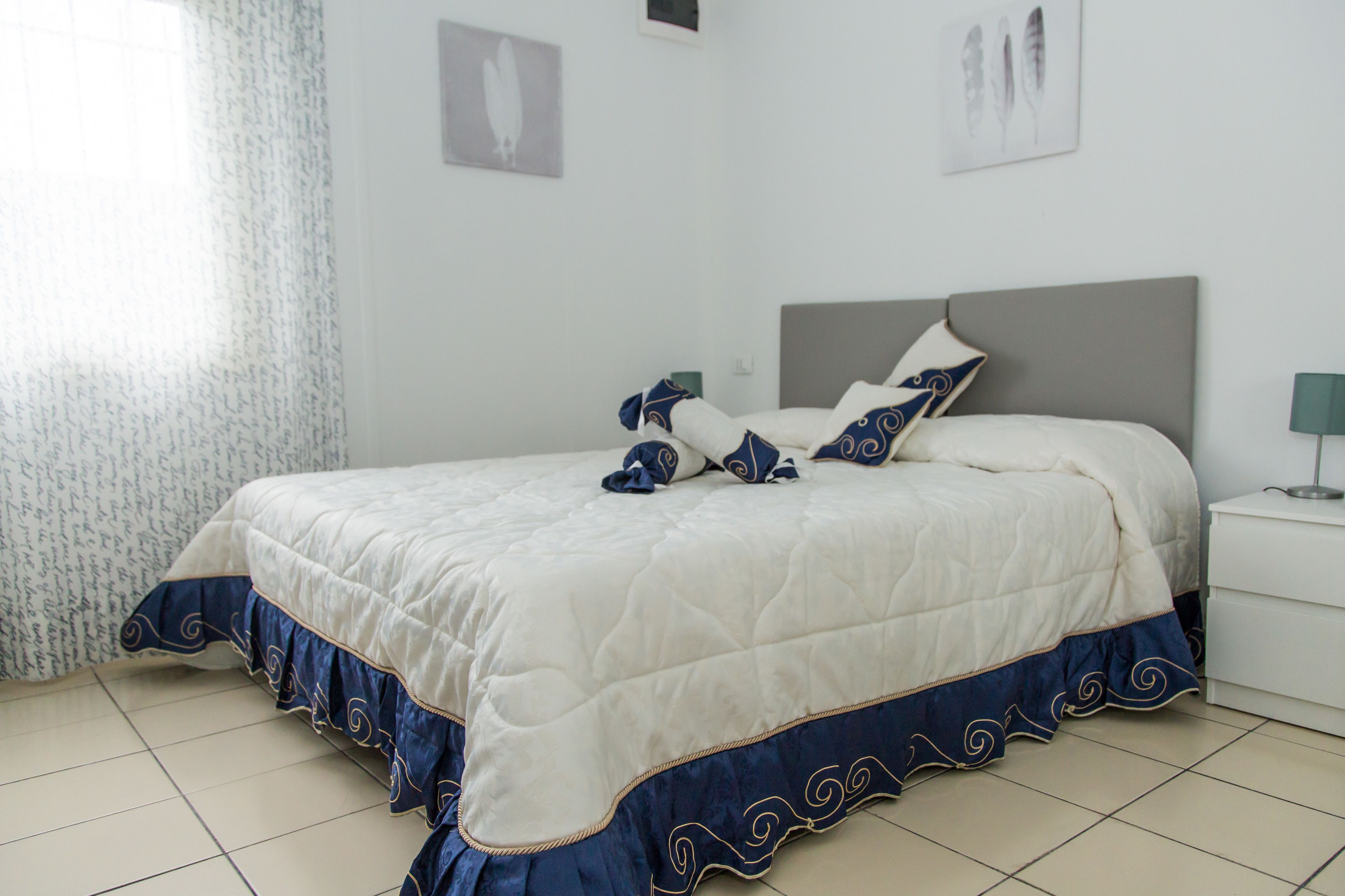 Una cama bien hecha con una colcha blanca y azul, un cabecero gris y una mesita de noche con una lámpara.