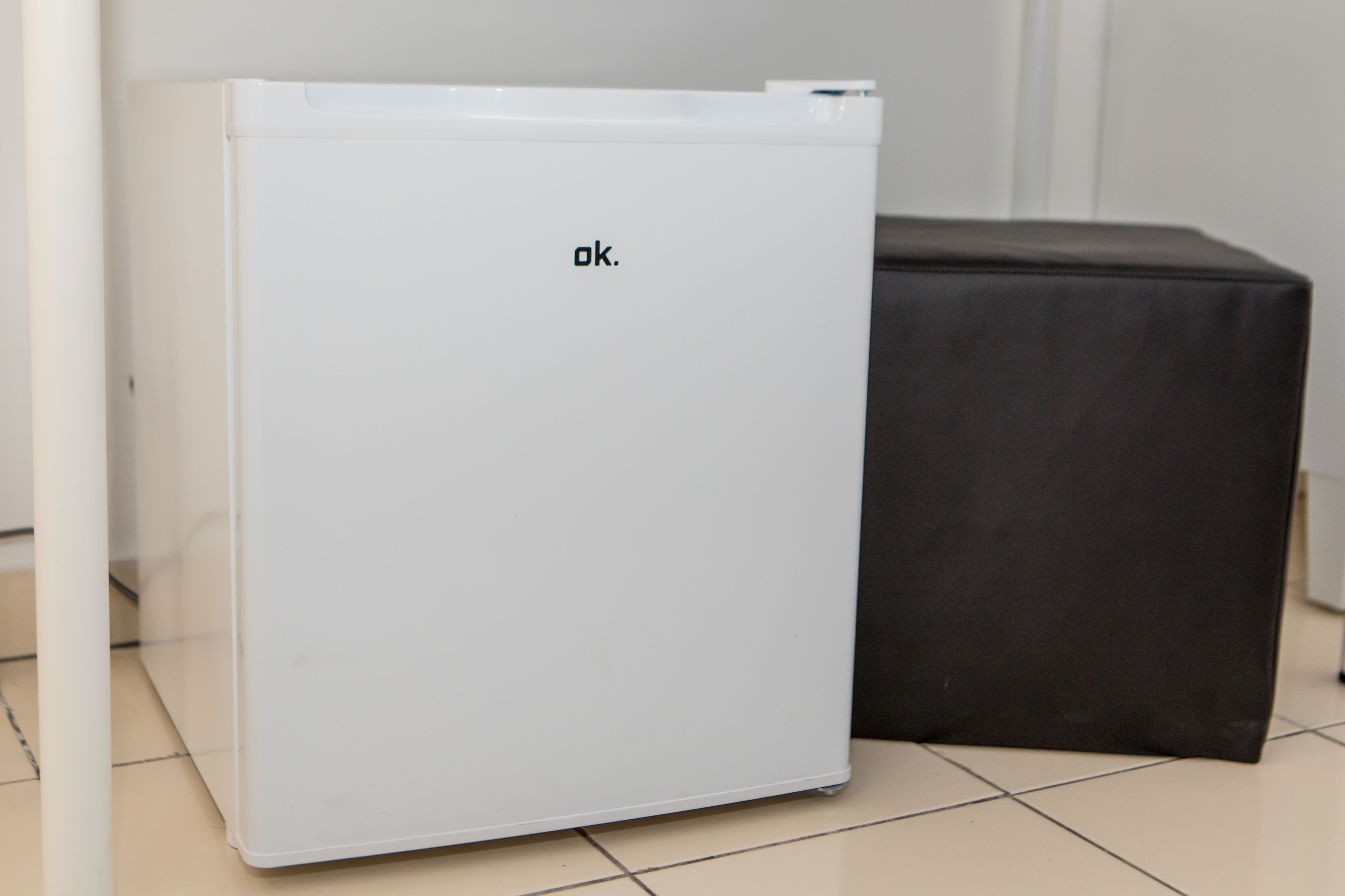 A white refrigerator with the brand name "ok." next to a black object, possibly a trash bin, in a room with tiled flooring.