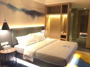A hotel room with a large bed, bedside table, lamp, and a wall-mounted shelf.