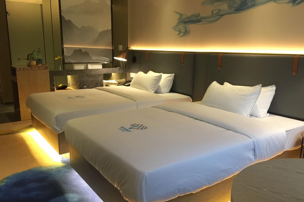 Une chambre d’hôtel avec deux lits, une petite table, un miroir et un mur orné d’un tableau représentant une montagne.