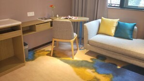 A small, neatly arranged room with a desk, chair, and sofa. There is a window with curtains, a small table with a vase and a magazine, and a carpet with a colorful pattern. - Lifu Hotel Shachong Metro Branch (Guangzhou)