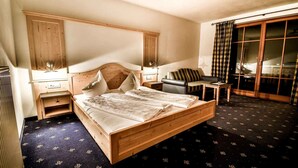 A hotel room with a wooden bed, a nightstand, a small table, a sofa, and a large window with curtains. - Hotel Vermoi (Laces)