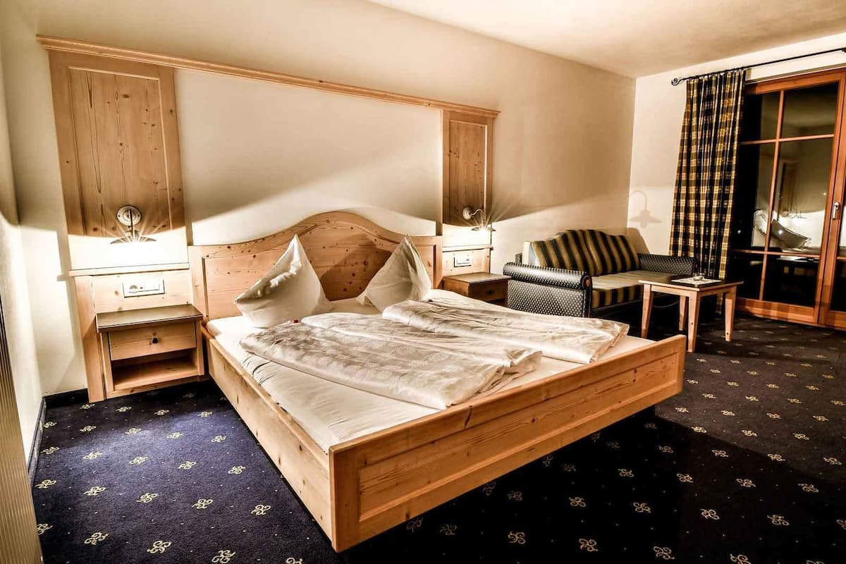Una habitación de hotel con una cama de madera, una mesita de noche, una pequeña mesa, un sofá y un ventanal con cortinas.