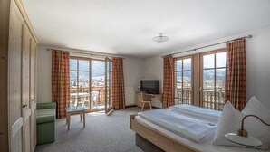 A bedroom with a bed, a television, a desk, and a chair. There are large windows with curtains, and a balcony with a view of the city. - Hotel Vermoi (Laces)