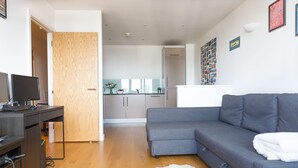 A modern living room with a grey sofa, a wooden door, a desk with a computer, and a kitchen in the background. - Modern apartment with great views (London)