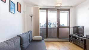 A modern living room with a grey sofa, a flat-screen TV on a wooden stand, and a city view through large sliding glass doors. - Modern apartment with great views (London)