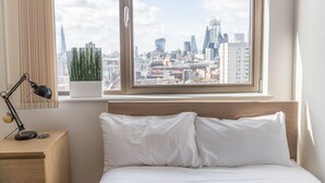 A hotel room with a bed, a desk, a lamp, a potted plant, and a view of a city skyline. - Modern apartment with great views (London)
