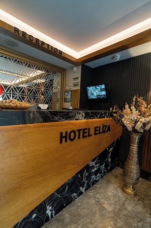 Mostrador de recepción de hotel con encimera de madera, cartel de recepción, cámara de seguridad, pantalla de televisión y un jarrón decorativo con flores.